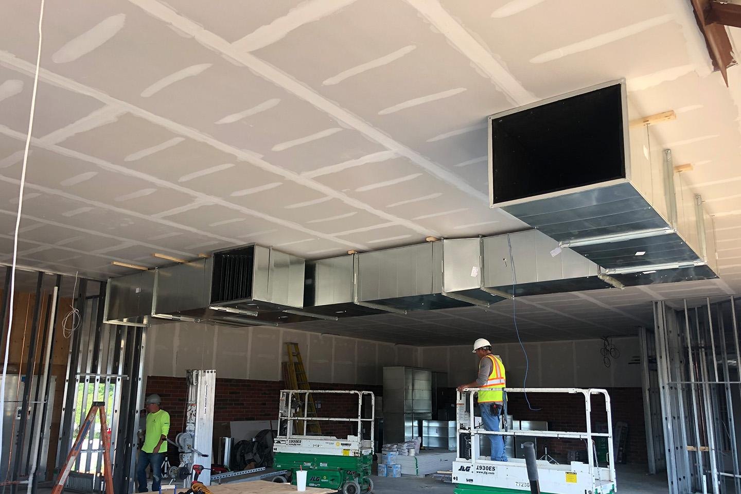 Worker on a lift beneath newly installed drywall and commercial ventilation ductwork